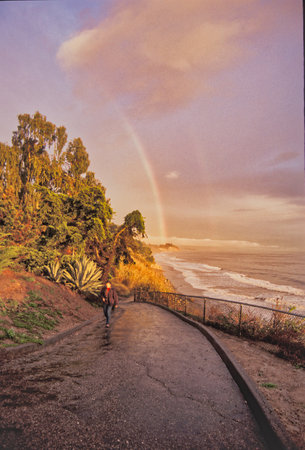 Woman walking on the path to the sea with rainbow in the skyの写真素材