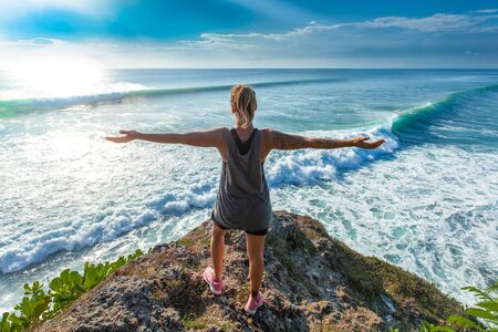 Surfer girl opening her arms standing on top of the high cliff in front of a breathtaking seascape,...