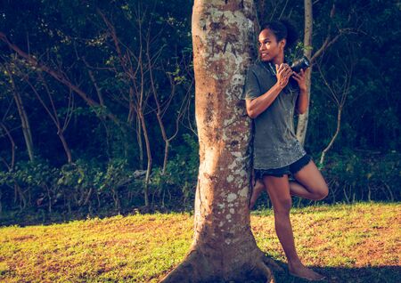Afro american young girl hiding with dslr camera behind the old big tree in the parkの写真素材