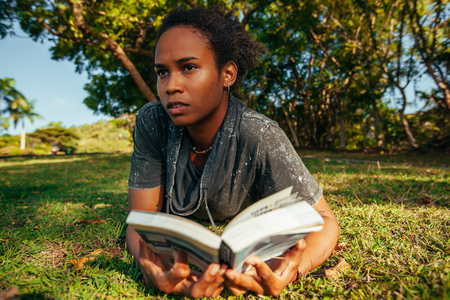 Beautiful young woman with afro hairstyle and book in her hands lying on the grass in the park on sunny summer dayの写真素材