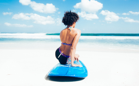 Surfer girl with afro hairstyle sitting on blue longboard on the white sand beach in front of a breathtaking seascape, looking at waves at Dreamland beach, Bali, Indonesiaの写真素材