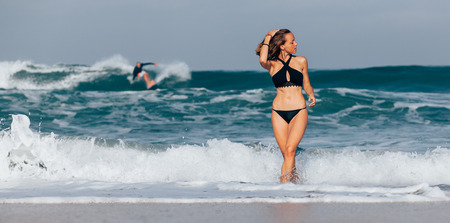 Beautiful girl in black swimwear coming out of the ocean with surfer riding the wave in the backgroundの写真素材
