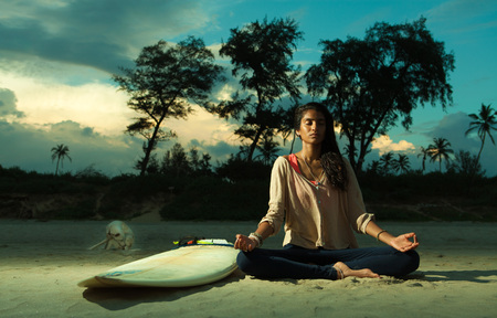 Indian surfer girl meditating in lotus pose on the beach at sunset next to surfboard and dog, eyes...