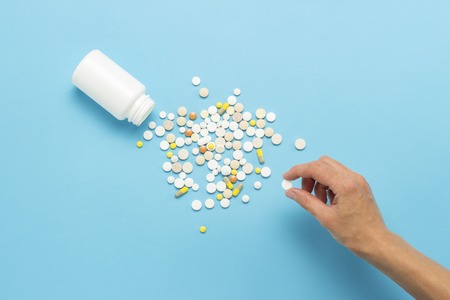 Female hand takes a pill from a pile of scattered pills and a white bottle on a blue background. Concept of taking drugs in case of illness, pharmacetiya, medicine. Flat lay, top view.の写真素材