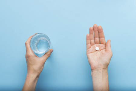 Female hand is holding a pill in the palm of a hand while the second hand is holding a glass of water on a blue background. Concept of taking drugs in case of illness and disease. Flat lay, top view.の写真素材