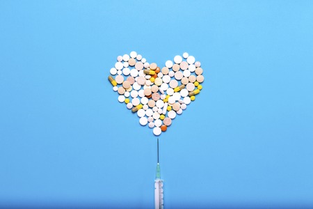 Pills in the shape of a heart and a stuck syringe on a blue background. Concept of the pharmaceutical industry, medicine, treatment and recovery after cardiovascular diseases. Flat lay, top view.の写真素材