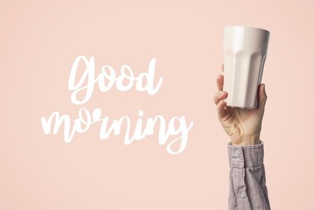 Female hand in clothes holding a purple cup with hot coffee or tea on a pink background. Added text Good morning. Breakfast concept with hot coffee or tea.の写真素材