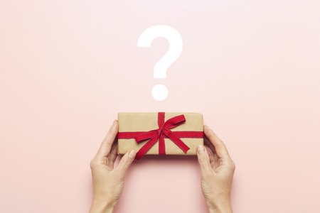 Female hands holding a gift box on a pink background with a question mark. Surprise, waiting for a gift for the holidays, birthday, christmas, wedding. Flat lay, top view.の写真素材