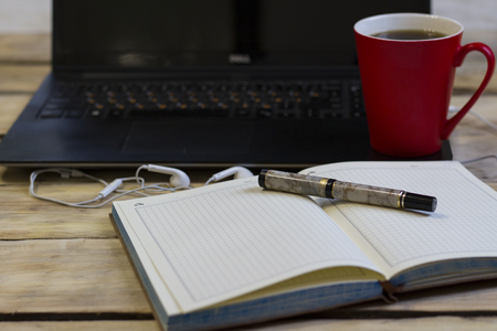 On the wooden table is a red cup with a coffee on the laptop. Headphones, notebook and pen. Pen in focus.の写真素材