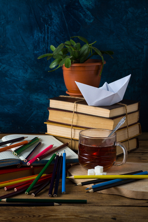 Pile of Books, A Cup of Hot Tea, A Paper Boat, An Open Diary with Color Pencils on a Dark Blue Background.の写真素材