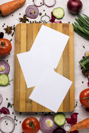 Fresh farm vegetables around a cutting board and a notebook with copy space. Tomatoes, Red onion rings, Spices, Cucumber, Carrots. cooking scene.の写真素材