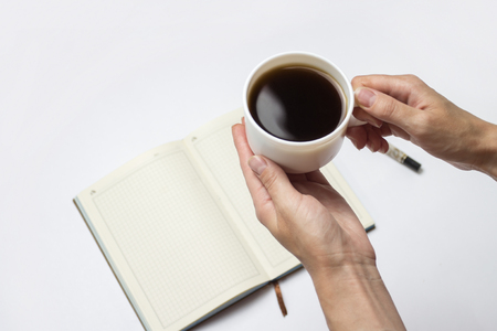 Female hands hold a cup of black coffee and an open diary with clean pages, a pen on a light background. Minimalist style and creative concepts. Flat lay, top view.の写真素材