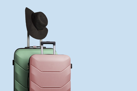 Two plastic suitcases on wheels and a wide-brimmed hat on the handle of a large suitcase against a blue background. Travel concept, vacation trip, visit to relatives.の写真素材
