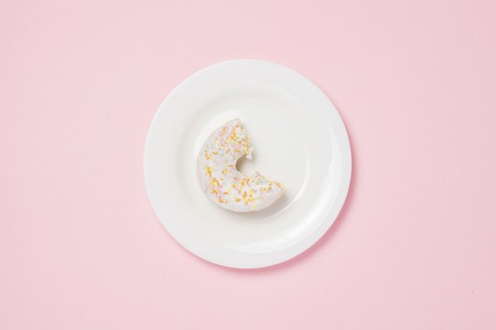 White plate with bitten off Fresh tasty sweet donut on a pink background. Bakery concept, fresh pastries, delicious breakfast, fast food, coffee shop. Flat lay, top viewの写真素材