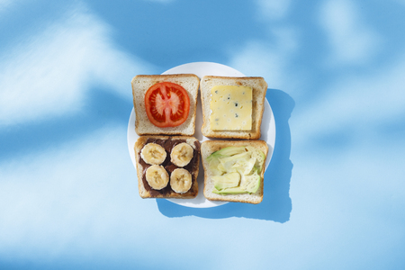 Sandwiches with cheese, tomato, banana and avocado on a white plate, blue background. Concept of healthy eating, breakfast at the hotel, diet. Natural lighting, hard light. Flat lay, top viewの写真素材