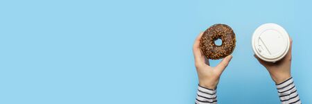 Female hands hold a donut and a paper cup with coffee on a blue background. Concept confectionery...