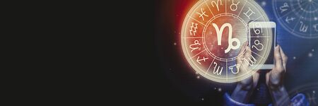 Female hands are holding a phone and an astrological circle with the zodiac sign Capricorn on a background of the starry sky. Mobile Application for the horoscope. Banner. Copy space.の写真素材