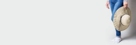 Woman holds in her hands a summer hat with wide brim on a light background. Only legs are visible. Banner.の写真素材