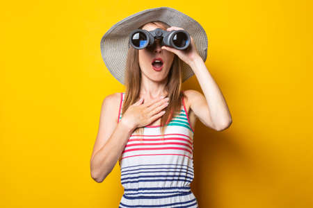 Young woman in a hat and a striped dress holds binoculars on a yellow background.の写真素材
