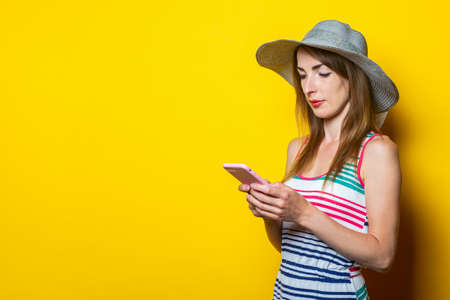 Serious young girl looks at the phone, in a hat and in a striped dress posing on a yellow background
