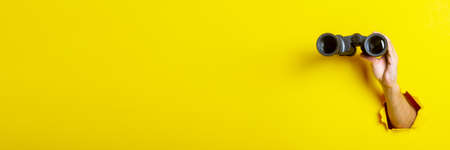 Female hand holds black binoculars on yellow background, travel, find and search concept. Banner.