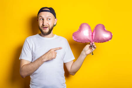 Cheerful bearded young man points finger at air pink balloons on yellow backgroundの写真素材