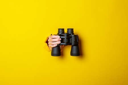 Female hand holds black binoculars on a yellow background. Journey, find and search concept.の写真素材