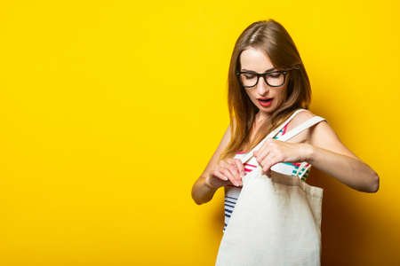 Surprised young woman looking into linen bag with purchases on yellow backgroundの写真素材