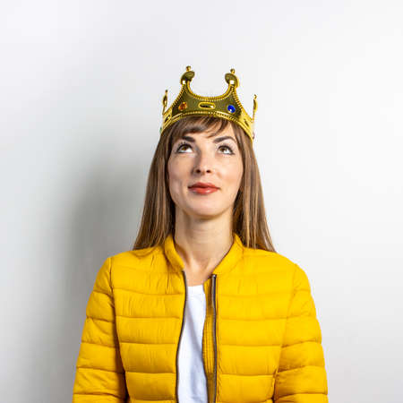 Young woman in a yellow jacket and a gold crown on a light background. Emotion laughter, surprise,...