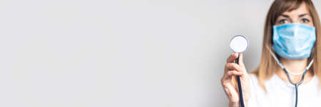 Young woman in a medical mask holds a stethoscope on a light background. Banner. Concept doctor,...