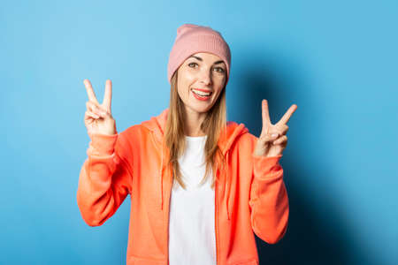 Beautiful young woman with a smile in a hoody and hat makes a gesture with her hands on a blue...