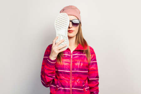 Young woman with glasses, hat and pink sports jacket with serious face covers her face with one sneaker on a white background with copy space. Concept modern trendy style. Face expression. Banner.の写真素材