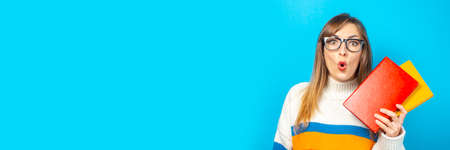 Young woman with a surprised face holds books in her hands on a blue background. Concept of...