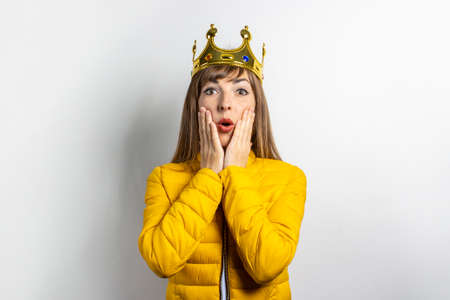 Young woman in a yellow jacket and a crown on her head with a surprised face on a light background. Emotion laughter, surprise, kiss. The concept of a queen, luck, the best, win, victory. Banner.の写真素材