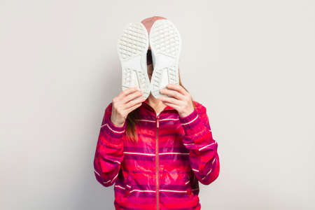 Young woman with glasses, hat and pink sports jacket with serious face covers her face with sneakers...