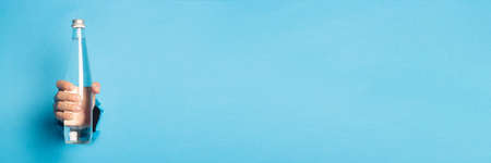 Man's hand holding a glass bottle with water on a bright blue background. Banner