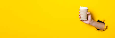 Man's hand holding a paper cup of coffee with the lid closed on a bright yellow background. Banner
