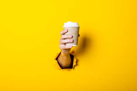 Man's hand holds a paper cup with coffee closed by a bump on a bright yellow background.の写真素材