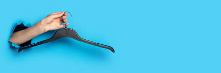 Female hand on a blue background holding a black plastic clothes hanger. Bannerの写真素材