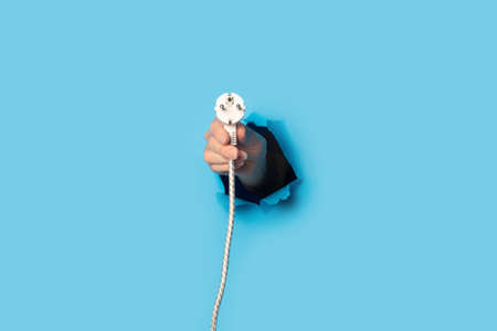Female hand holding a plug with a wire on a bright blue background.