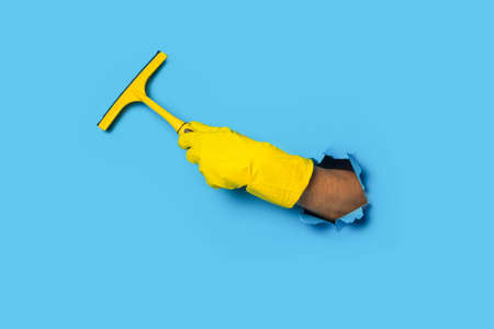 Hand in yellow gloves holding a rake for cleaning windows on a bright blue background.の写真素材