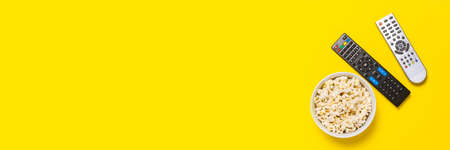 Two remotes from the TV, TV tuner and a bowl of popcorn on a yellow background. Concept series, film, sports. Banner. Flat lay, top view.の写真素材