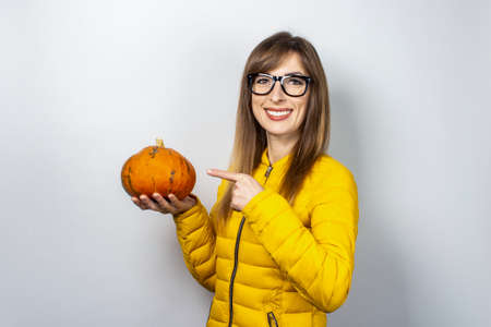 young girl in a yellow jacket holds a pumpkin and points a finger at it on a light background. Halloween concept, autumn, celebration.の写真素材