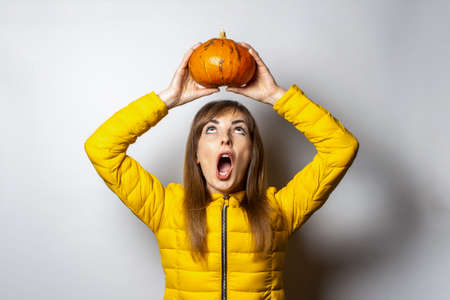 young girl holding a pumpkin over his head and looking at her with a frightened face on a light background. Halloween concept, autumn, celebration.の写真素材
