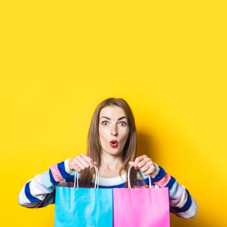 Young woman in sweater holds shopping bags with purchases on yellow background.