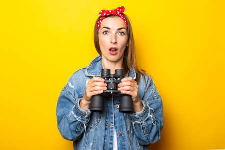 Young woman with a surprised face in jeans and a raft on her head holds binoculars in her hands on a yellow background. Banner.の写真素材