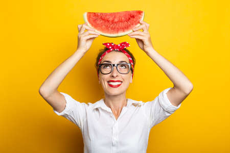 Young woman with a smile in a red headband and glasses holds a watermelon above her head on a yellow...
