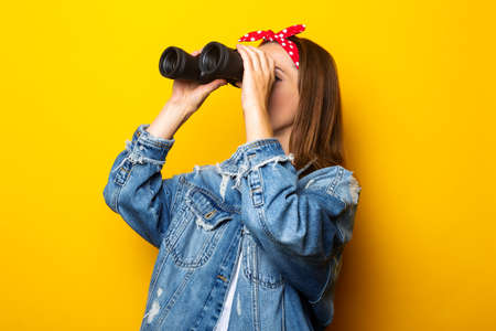 Young woman in a denim jacket and a raft on her head looks through binoculars on a yellow...