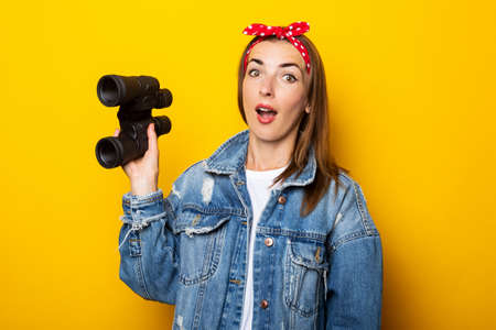 Young woman with a surprised face in jeans and a raft on her head holds binoculars in her hands on a...