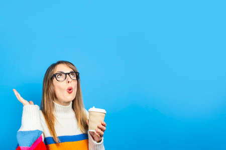 young girl in a knitted sweater holds a cardboard cup with coffee and shows emotions, surprise on a...